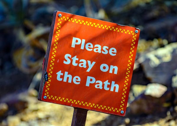 A red sign affixed to a metal post in a rocky outdoor area that says "Please Stay on the Path"